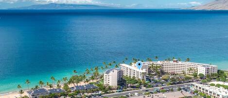 A coastal hotel with a central building, a parking lot, and a beach area.