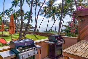 An outdoor patio with two grills, a wooden table, and a view of the ocean with palm trees. - Wailea Elua 803 - Ocean Views, Modern Renovation, Central AC, Wi-fi, BBQ Grill (Kihei)