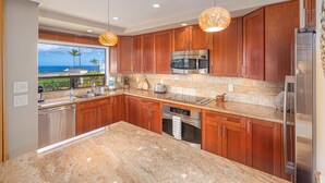 A modern kitchen with granite countertops, stainless steel appliances, wooden cabinets, and a view of the ocean. - Dramatic Ocean Views, Wailea Ekolu 407, Perfect for Couples, Renovated, Split AC (Kihei)