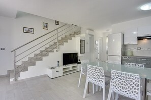 Ein modernes Apartment mit Treppe, einem Essbereich mit weißen Stühlen, einer Küche mit weißen Schränken und einem Flachbildfernseher auf einem weißen Schrank.