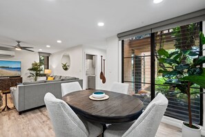 A modern living room with a dining table, grey sofas, a flat-screen TV, and a large window with blinds. - Wailea Ekahi 24B - Modern Garden Retreat w/ Lanai, Split AC & Stylish Renovation (Kihei)