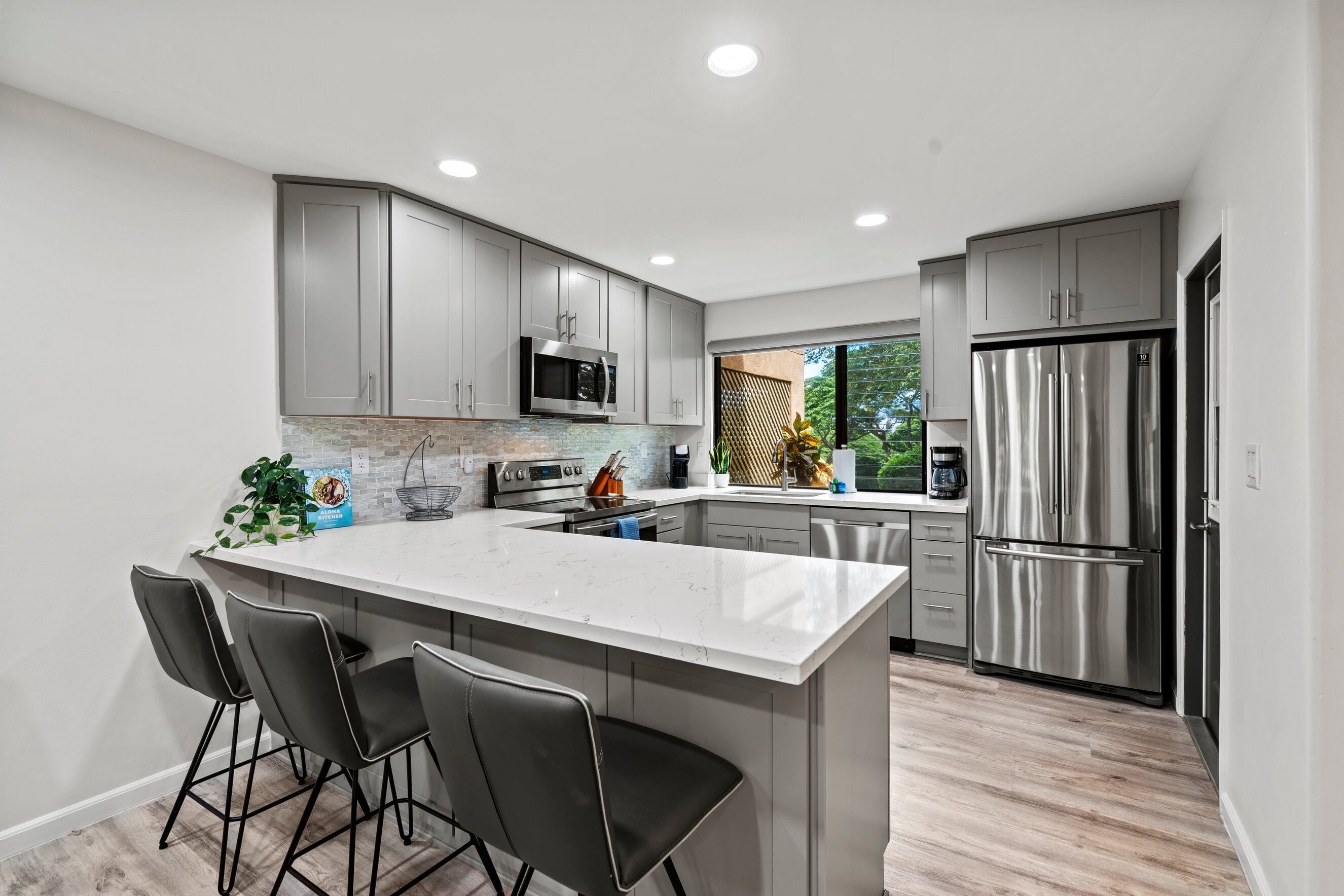 A modern kitchen with grey cabinets, stainless steel appliances, a marble island, and a view of greenery outside.