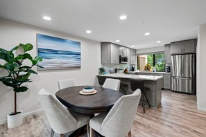 A modern kitchen with grey cabinets, stainless steel appliances, and a dining area with a round table and grey chairs. - Wailea Ekahi 24B - Modern Garden Retreat w/ Lanai, Split AC & Stylish Renovation (Kihei)