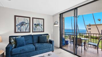 Un salón moderno con un sofá azul, una mesa de centro y un balcón con vistas al océano y palmeras.