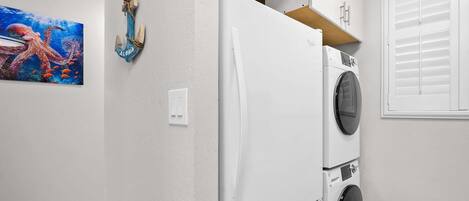 A laundry room with a white refrigerator, a washing machine, and a wooden shelf above the refrigerator.