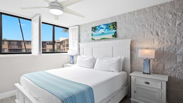 Un dormitorio con una cama grande, un ventilador de techo, una mesita de noche con una lámpara y un cuadro con motivos playeros en la pared.
