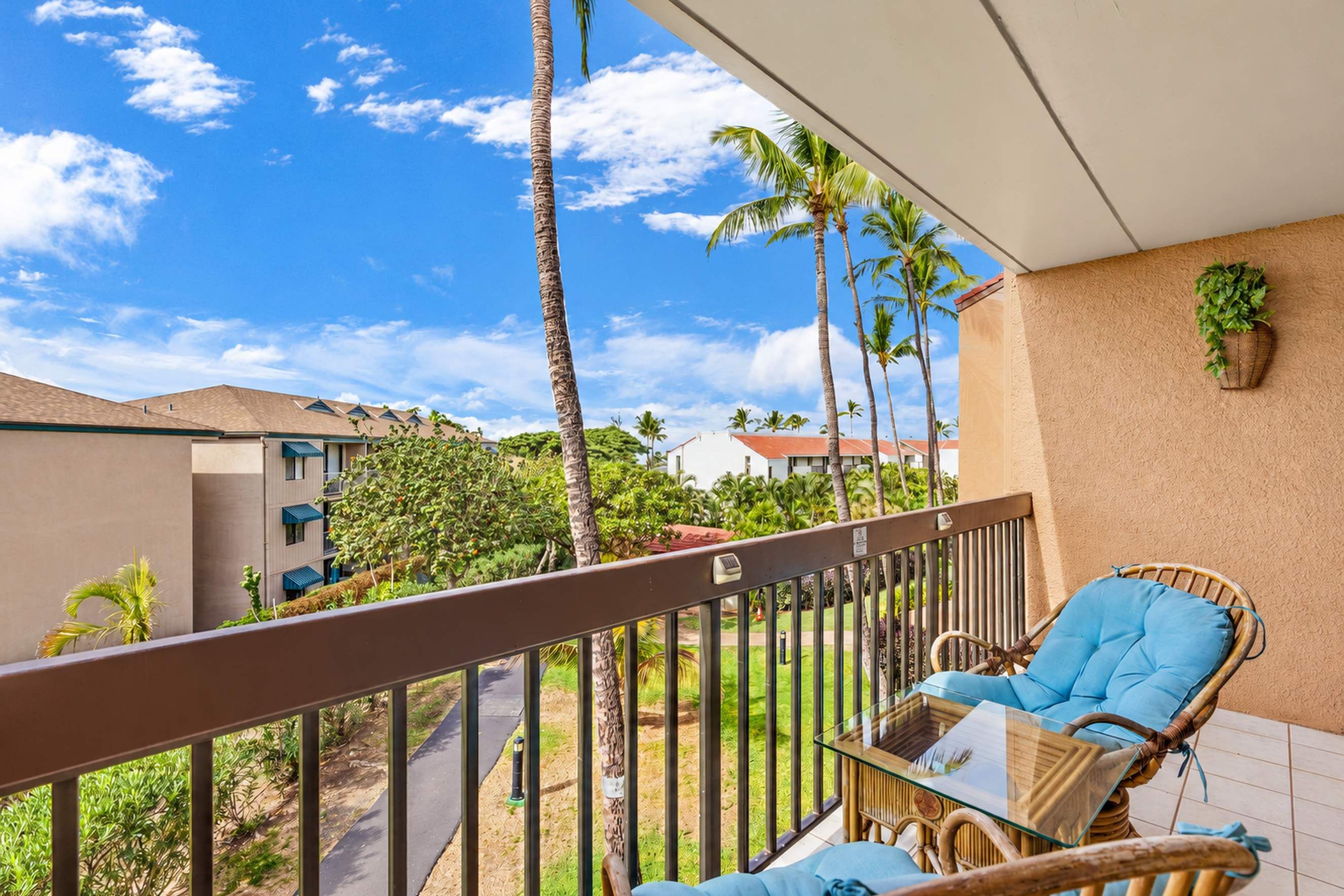 A balcony with a wicker chair and a glass table, overlooking a residential area with palm trees and houses.