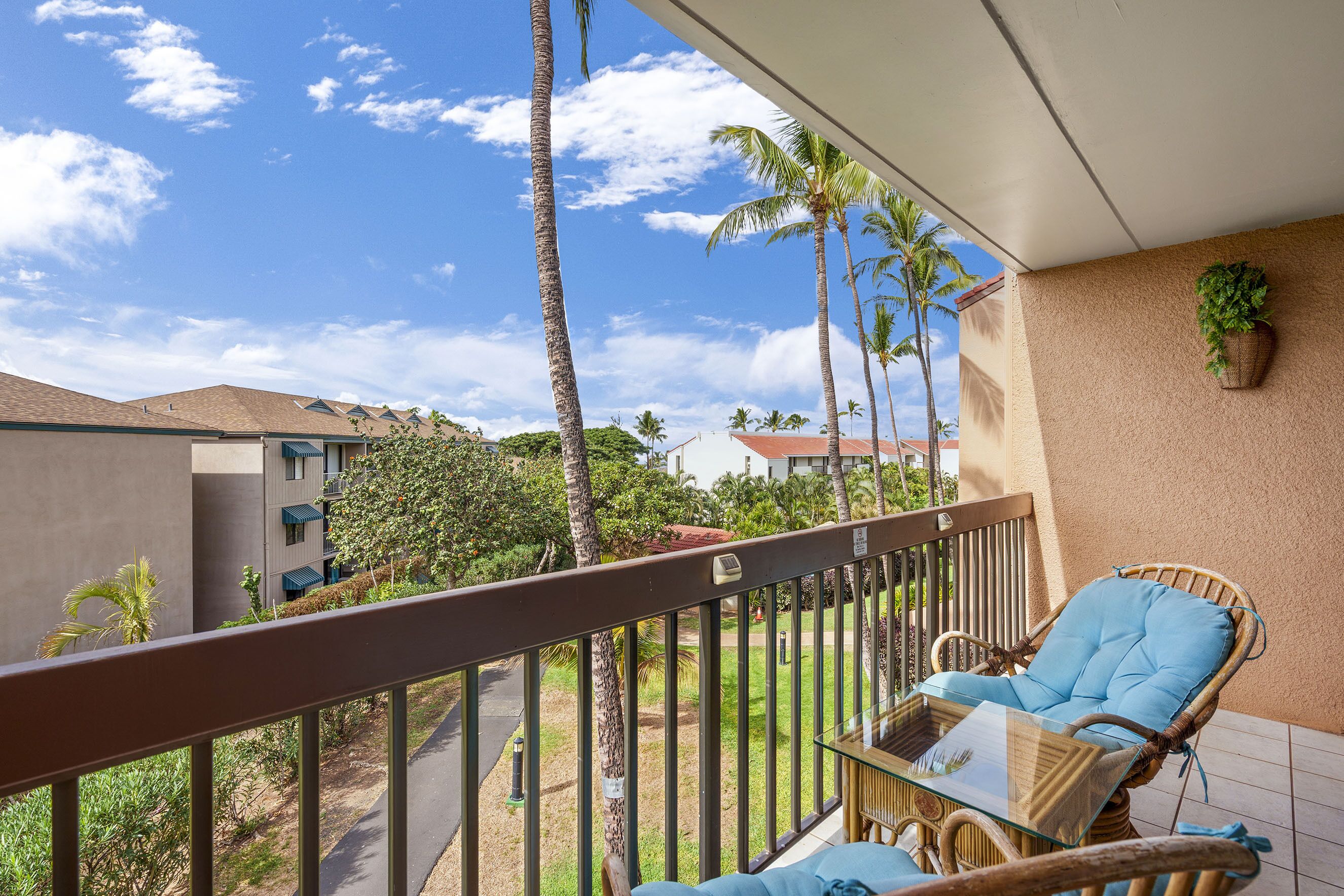 A balcony with wicker furniture, a glass table, and a view of palm trees and buildings.