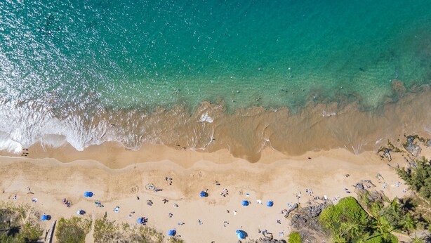 Vue aérienne d’une plage avec une eau turquoise et limpide, un rivage sablonneux et des parasols dispersés.