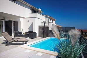 Een modern huis met een zwembad, tuinmeubilair en uitzicht op zee.