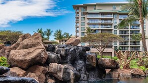 A hotel with a large rock formation and a pool in the foreground, with a palm tree and a building in the background. - Honua Kai Hokulani 934, Renovated, AC, Large Lanai, No Carpet, Amazing Views! (Lahaina)