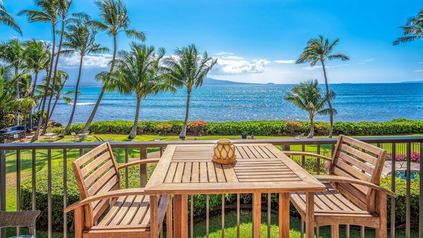 A wooden table and chairs on a balcony with a view of the ocean and palm trees.