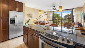 A modern kitchen with a stainless steel refrigerator, double oven, and a large island with a built-in stove. There are wooden cabinets, a ceiling fan, and a view of the outdoors through large windows. - Maui Vista 1-308 - Steps From the Beach, Renovated, Ocean Views, New Windows, AC (Kihei)