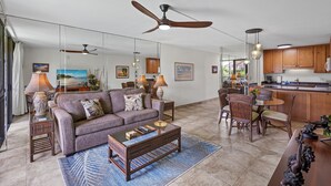 A spacious living room with a sofa, coffee table, and dining area with chairs. There is a ceiling fan, kitchen area with cabinets, and a large window. - Kamaole Sands 7-206 - Renovated, Courtyard Views, Steps to the Beach, AC (Kihei)