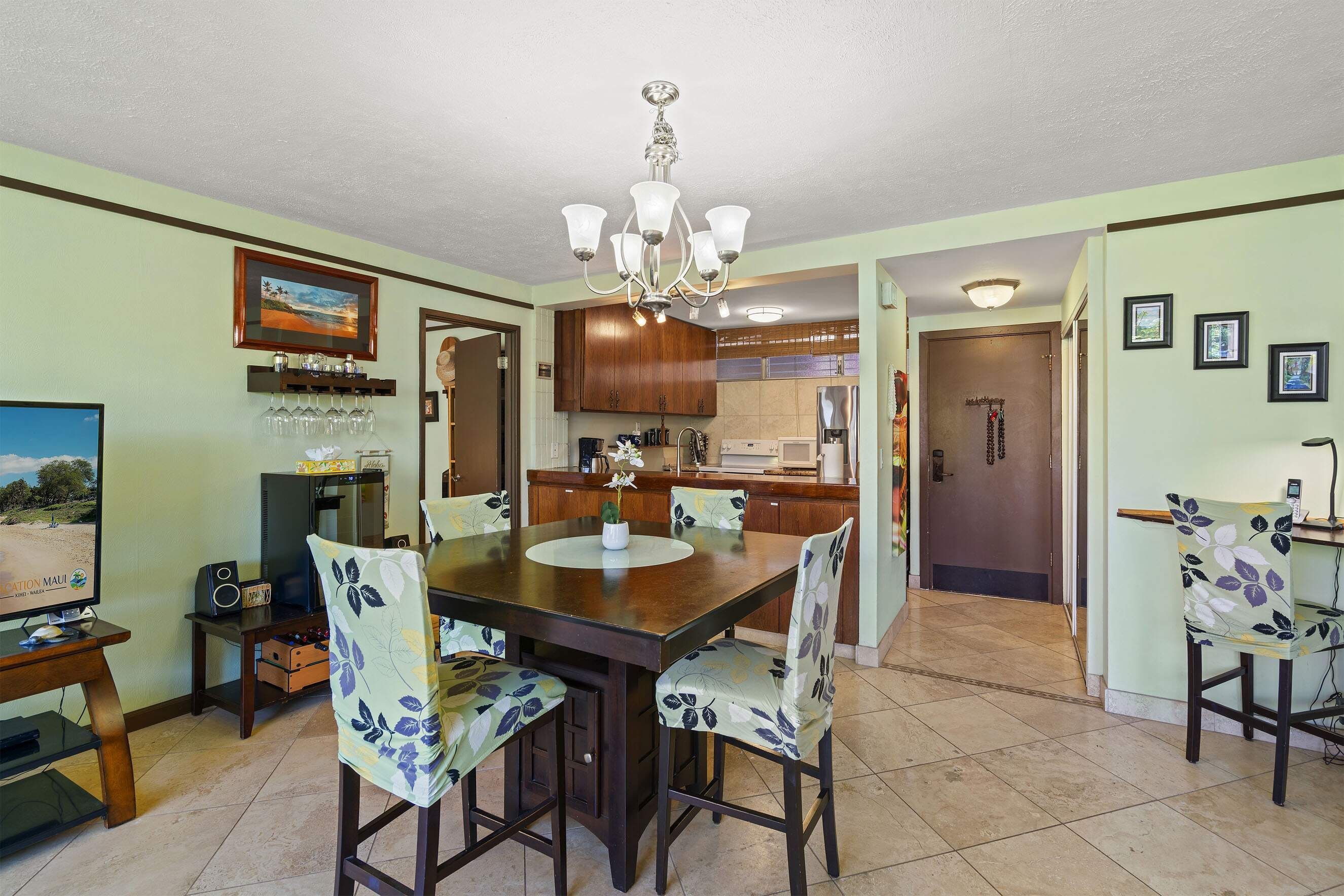 A dining area with a wooden table and chairs, a chandelier, and a kitchen in the background.