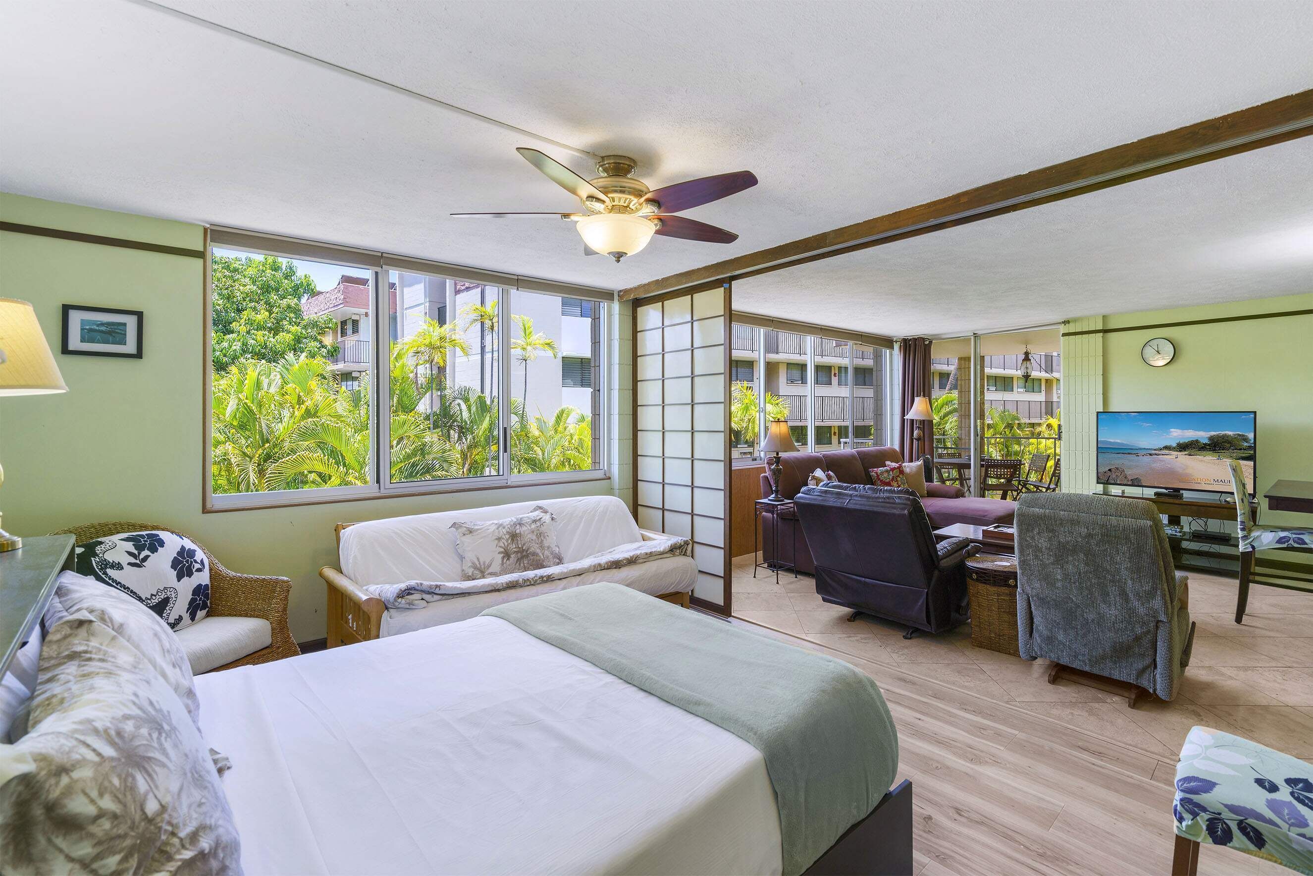 A room with a bed, a sofa, a chair, and a television. There are windows with a view of greenery outside.