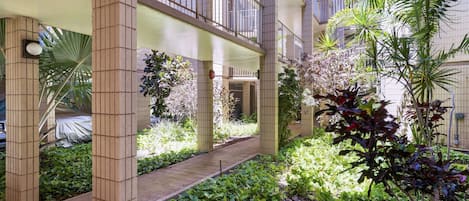 A covered outdoor area with a pathway, surrounded by greenery and a building with balconies.