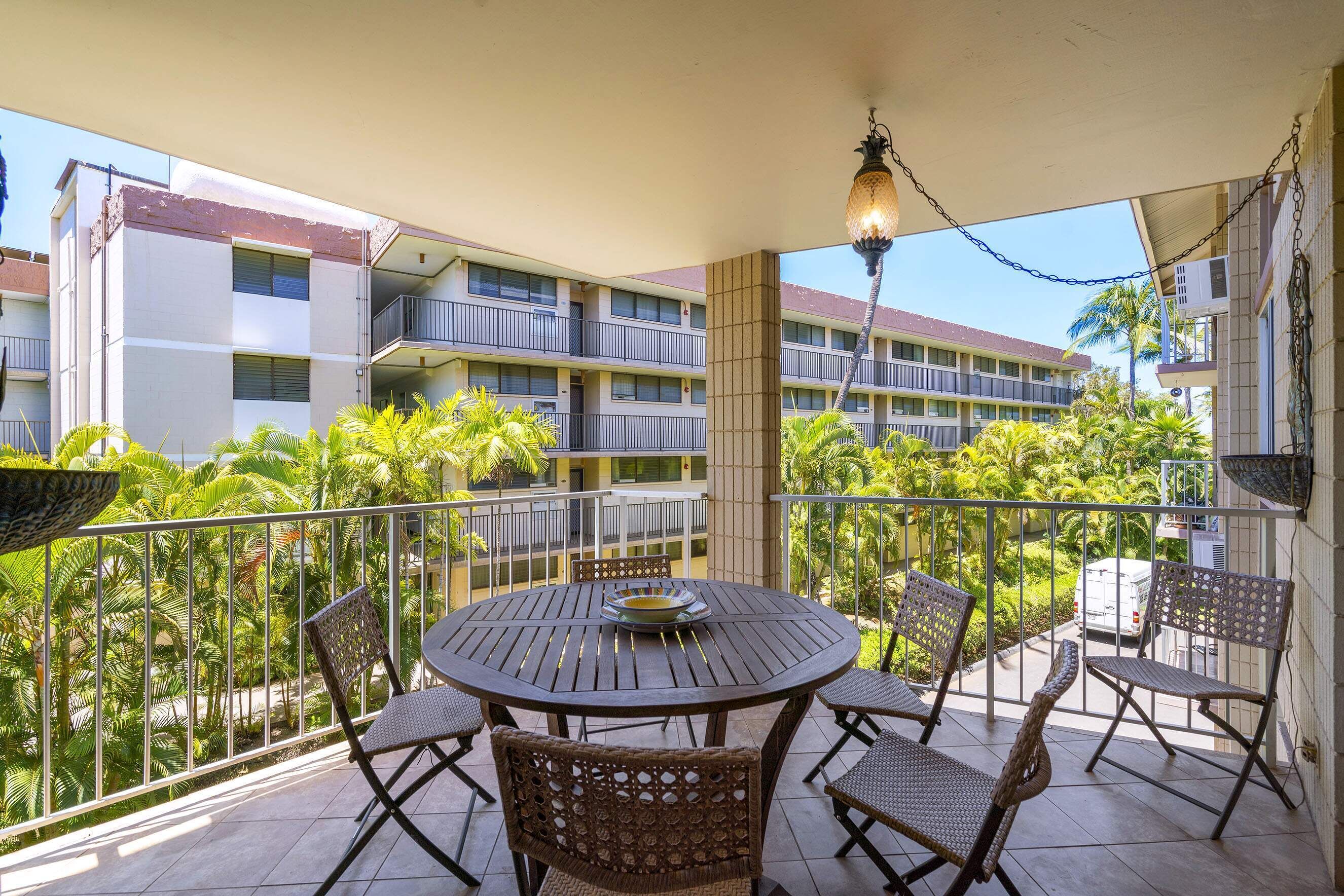 A balcony with a round table and chairs, surrounded by a metal railing, overlooking a building with multiple balconies and palm trees.
