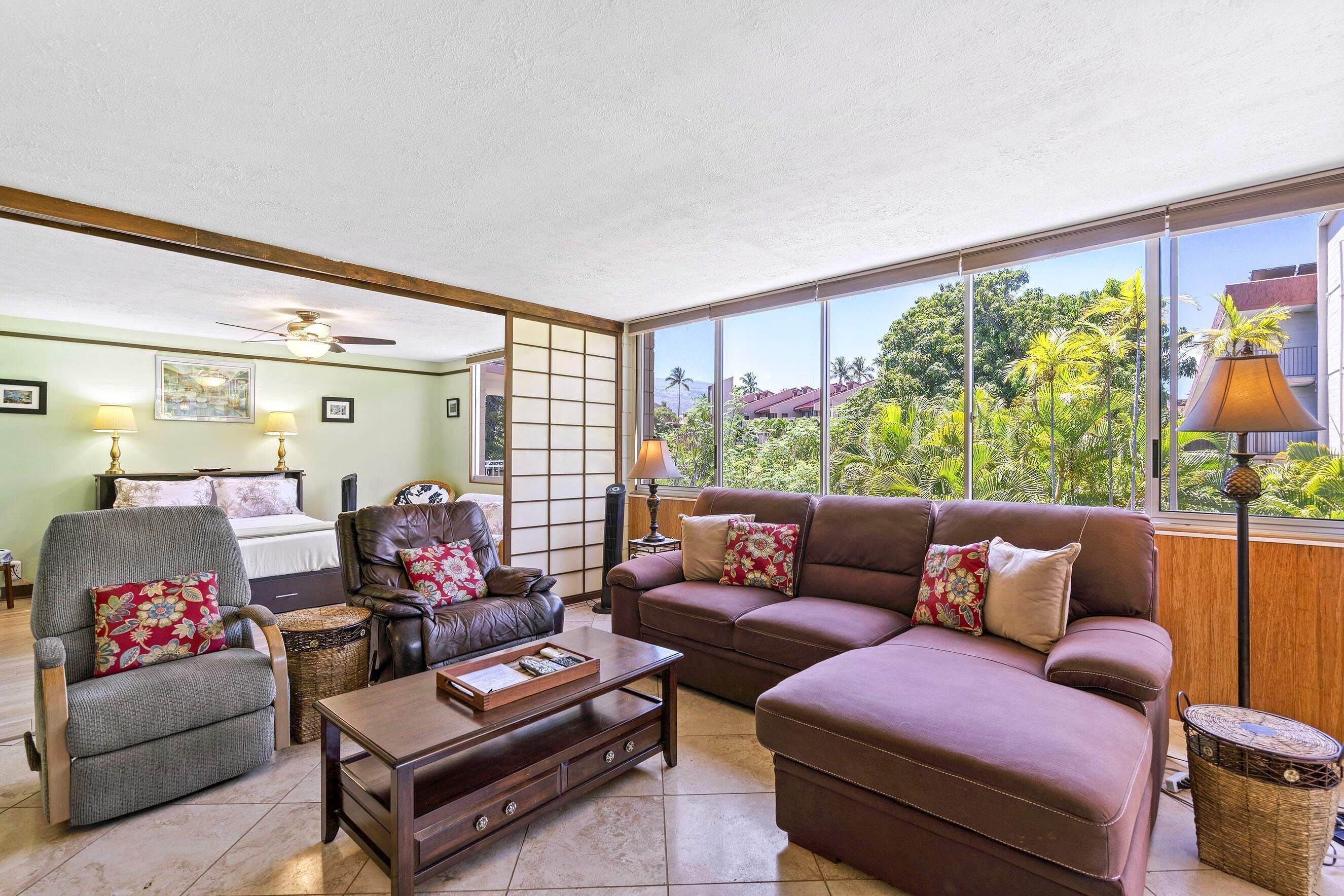 A living room with a sofa, armchair, and coffee table, a bed with a headboard, and a view of greenery through large windows.
