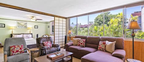 Una sala de estar con sofá, sillón y mesa de centro, cama con cabecera, y vista a la vegetación a través de amplias ventanas.