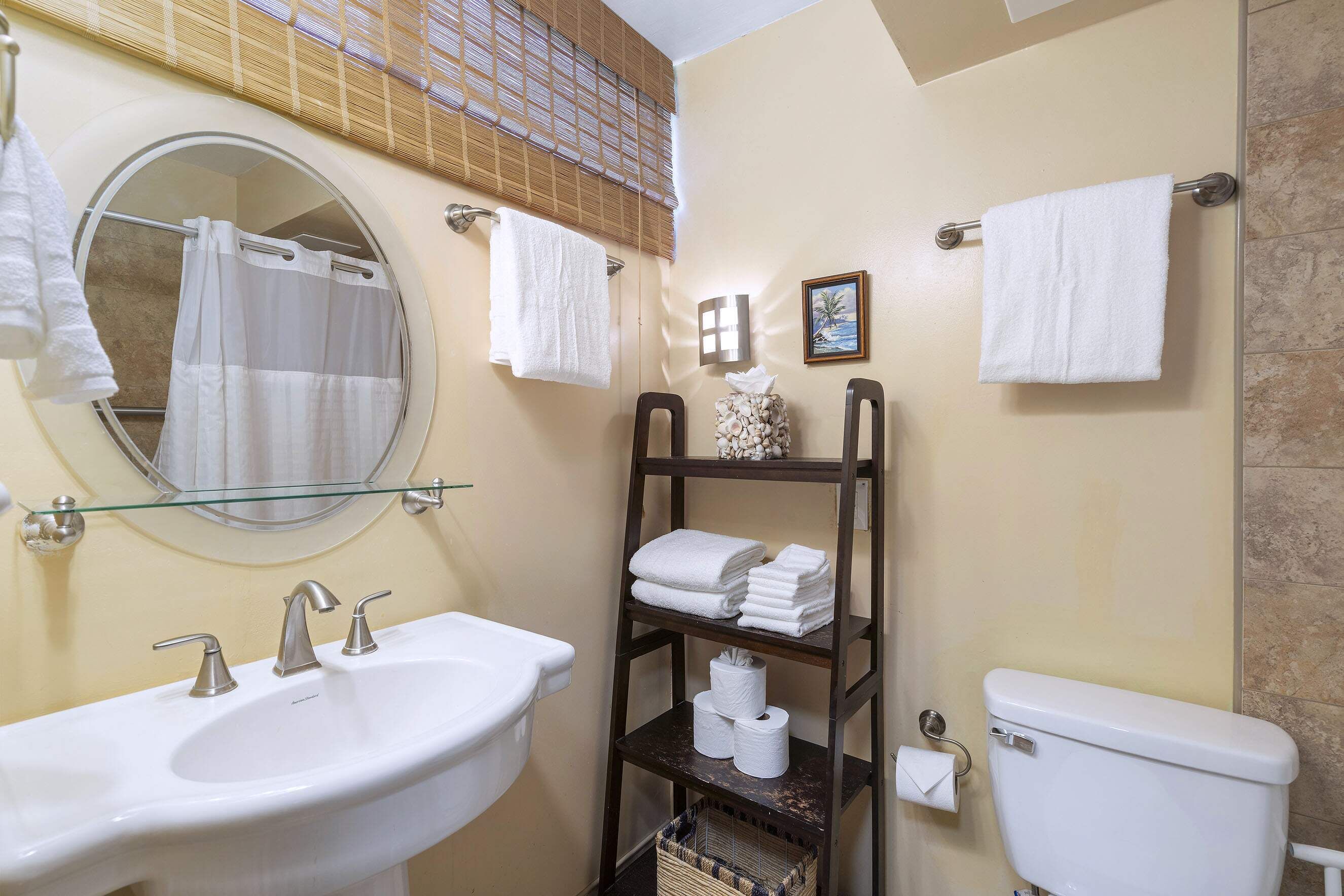 A bathroom with a round mirror, two sinks, a toilet, and a shelf with towels and toilet paper.