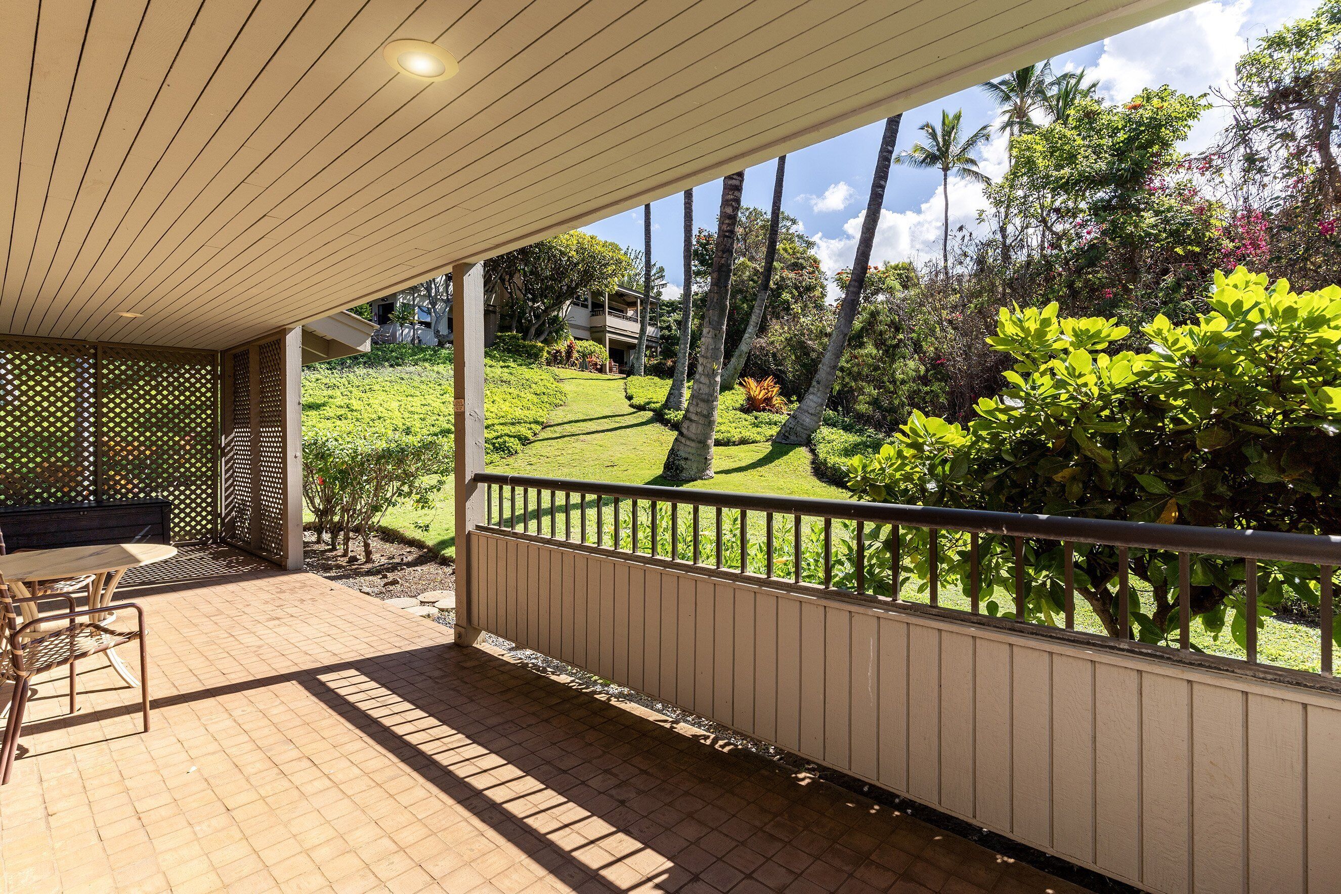 A covered patio area with a table and chairs, a view of a lush garden with palm trees and a building in the background.