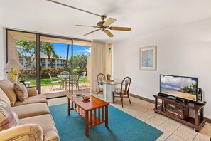 A living room with a sofa, coffee table, TV on a wooden stand, and a balcony with outdoor furniture. - Maui Vista 3-102 - Updated, Ground Level, Steps to the Pool, Walk to Everything! (Kihei)