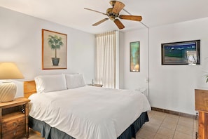 A hotel room with a bed, a ceiling fan, a window with curtains, a bedside table with a lamp, and artwork on the walls. - Maui Vista 3-102 - Updated, Ground Level, Steps to the Pool, Walk to Everything! (Kihei)