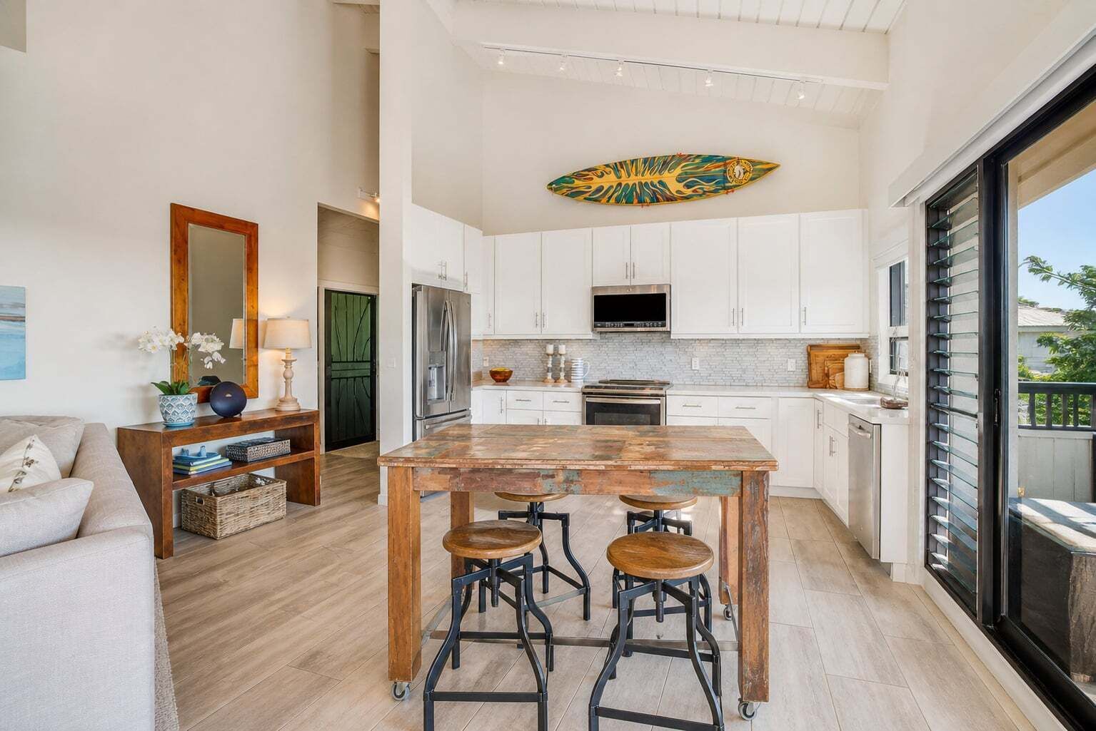 Une cuisine moderne avec des meubles blancs, un îlot central en bois doté de tabourets, une grande fenêtre et une décoration sur le thème du surf.