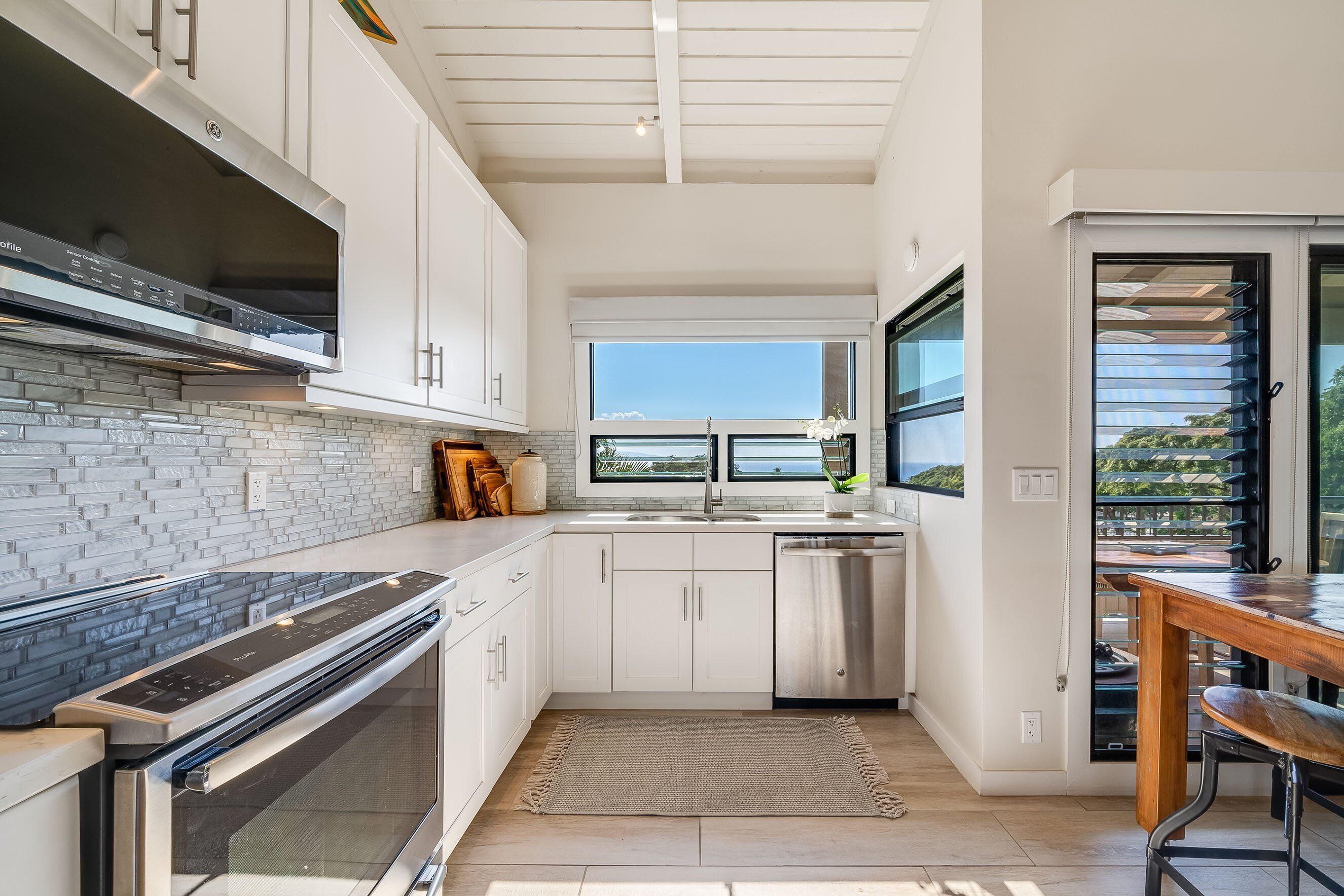 A modern kitchen with white cabinetry, a stainless steel range hood, a dishwasher, and a large window providing a view of the outdoors.
