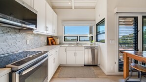 A modern kitchen with white cabinetry, a stainless steel range hood, a dishwasher, and a large window providing a view of the outdoors. - Extraordinary Molokini Views, Spectacular Renovation, New Split AC, Ocean Views! (Kihei)