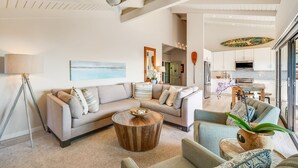 A spacious living room with a large sofa, a coffee table, and a dining area with a TV and kitchen in the background. - Extraordinary Molokini Views, Spectacular Renovation, New Split AC, Ocean Views! (Kihei)