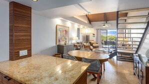 A spacious kitchen with granite countertops, a dining area with a large wooden table, and a view of the outdoors through a sliding glass door. - Kamaole Sands 10-403 - Stunning Renovation - Penthouse Level - Split AC - Look! (Kihei)
