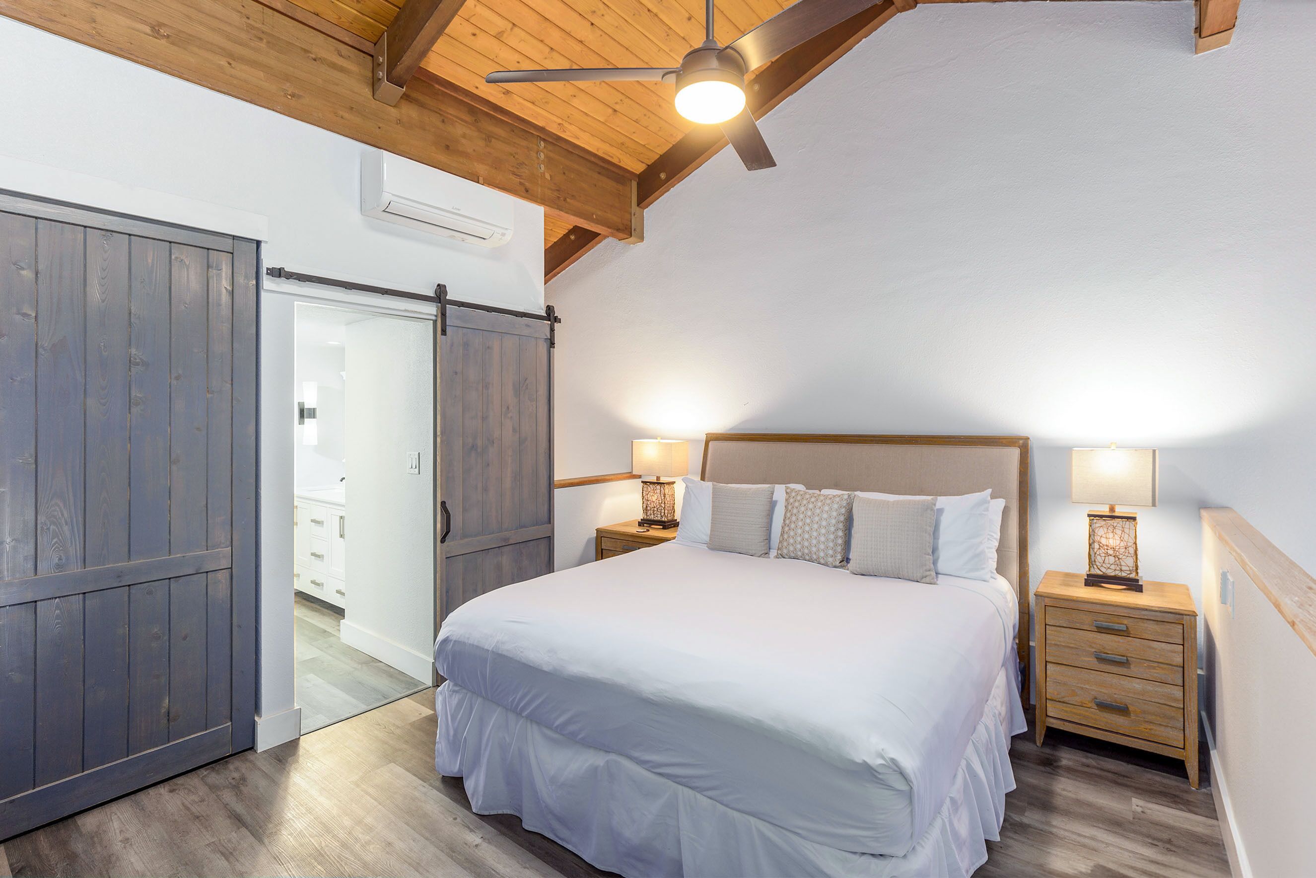 Un dormitorio con una cama grande, vigas de madera en el techo, un ventilador de techo y una mesita de noche con lámpara.