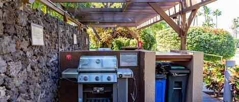An outdoor barbecue area with a stone wall, a grill, and a trash bin.