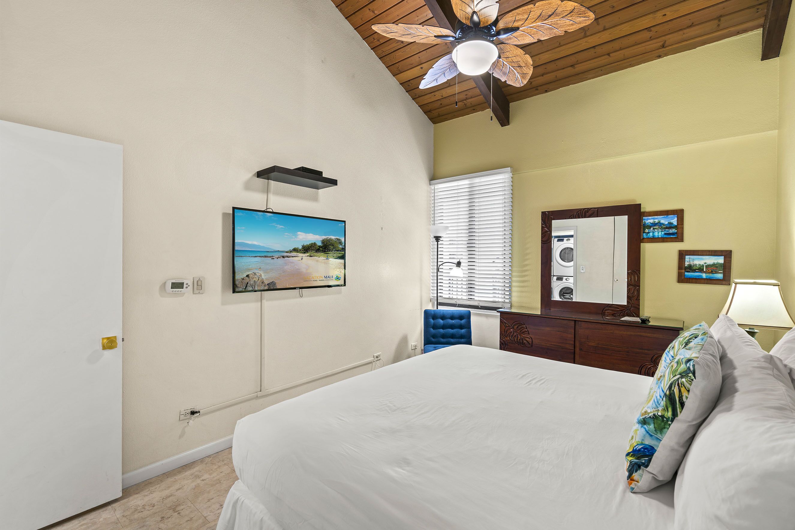 Une chambre à coucher comprenant un lit, un ventilateur de plafond, une télévision, une fenêtre avec des stores, un miroir et une commode.