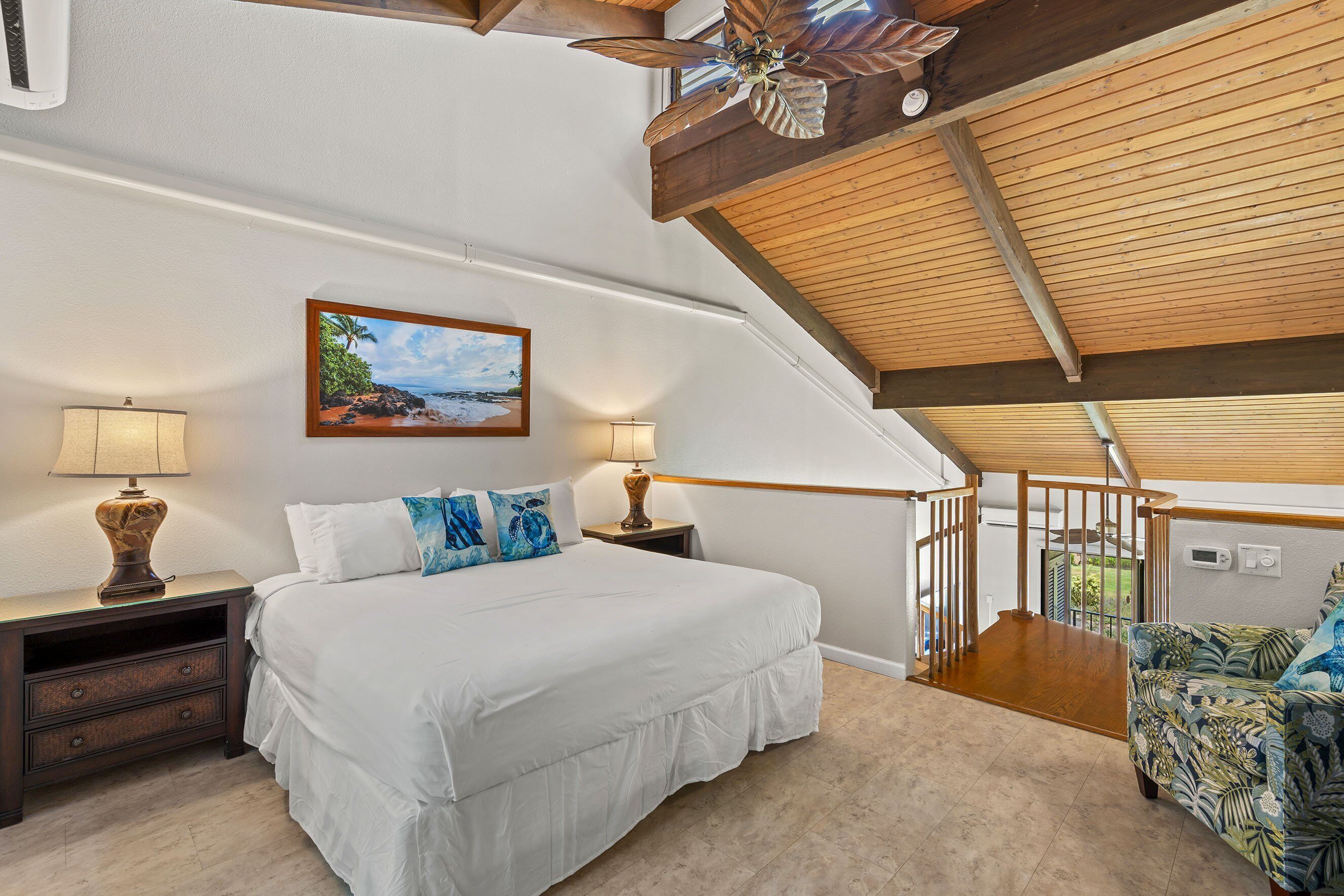 Ein Schlafzimmer mit einem großen Bett, zwei Nachttischen mit Lampen, einem Deckenventilator, einem Strandbild und einer Treppe, die in eine obere Etage führt.