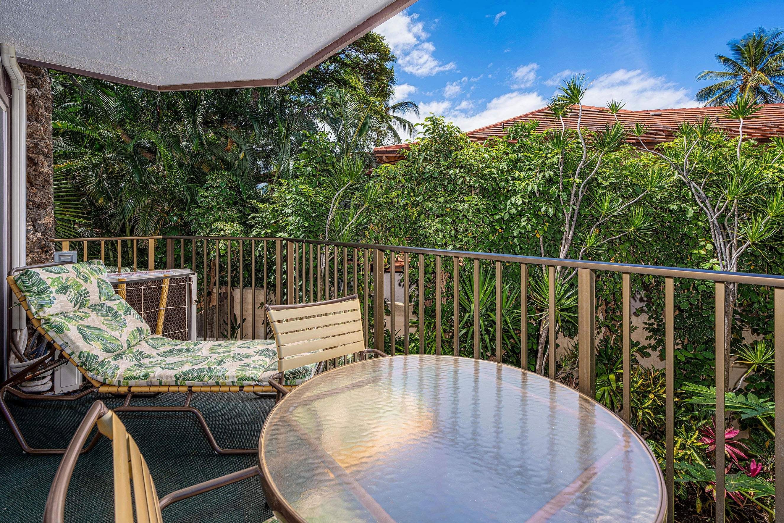 Un balcone con tavolo in vetro e due sedie, immerso nel verde e con cielo sereno.