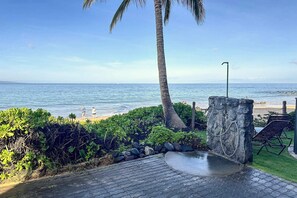 A paved pathway leading to a beach with a palm tree and a stone wall. - Hale Hui Kai 202 - Keawakapu Beach, Garden Views, Steps to the Beach, Split AC! (Kihei)