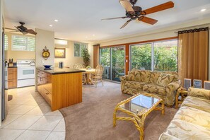 A spacious living area with a sofa, a coffee table, and a dining table. There is a kitchen area with a stove and cabinets. The room has large windows and a ceiling fan. - Hale Hui Kai 202 - Keawakapu Beach, Garden Views, Steps to the Beach, Split AC! (Kihei)