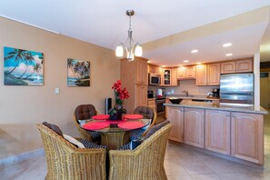 A modern kitchen with wooden cabinets, a stainless steel refrigerator, and a dining area with wicker chairs and a glass table. - Kamaole Sands 2-204 - Dramatic Ocean and Courtyard Views, 1bd / 2ba, AC, Wi-fi (Kihei)
