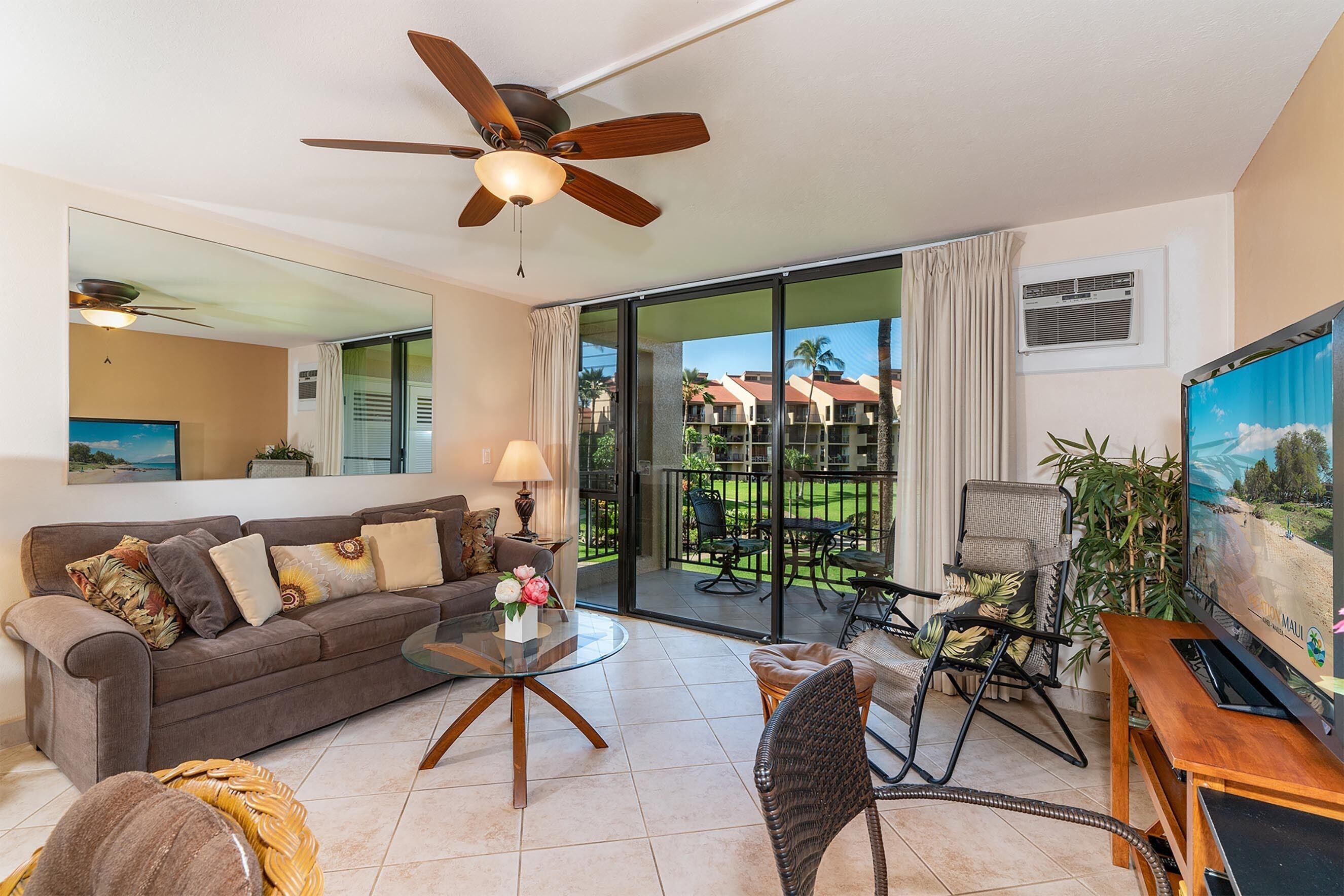 A spacious living room with a large sofa, a coffee table, and a TV. There's a balcony with outdoor furniture and a view of palm trees.