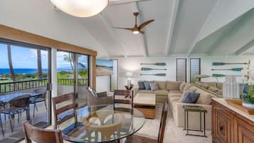 A spacious living area with a glass table, wooden chairs, a sofa, and a view of the ocean through large windows.