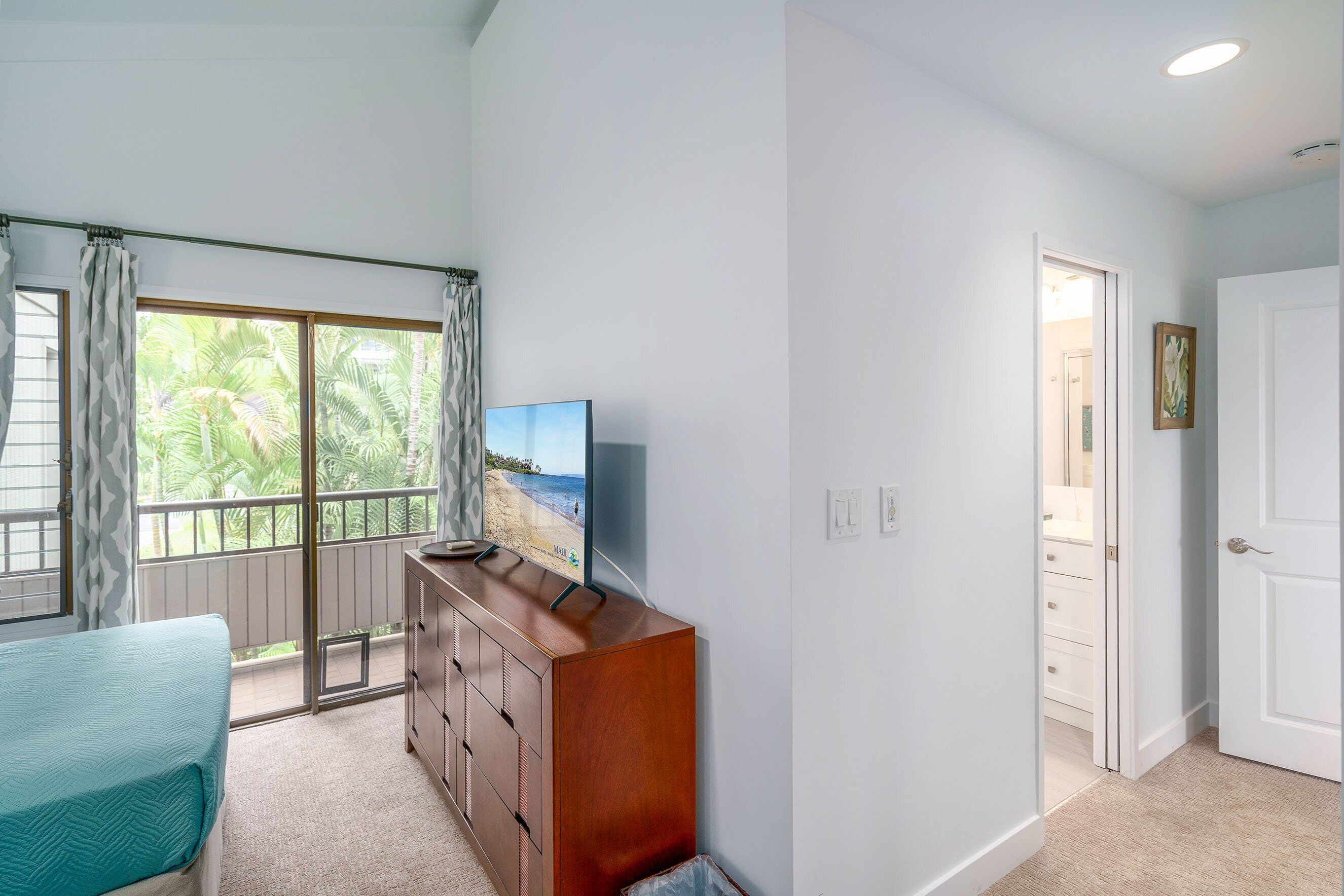 A bedroom with a bed, a dresser, a TV, and a balcony with a view of palm trees and the beach.