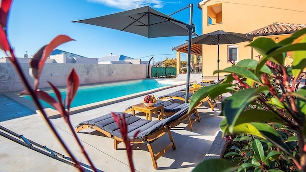 Un espace aménagé au bord de la piscine, avec des chaises longues et des parasols, un ciel d’un bleu éclatant et un bâtiment en arrière-plan.