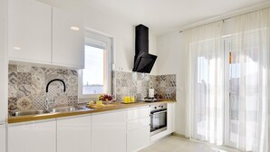 Une cuisine moderne avec des armoires blanches, un évier en acier inoxydable, une hotte noire et une fenêtre ornée de carreaux décoratifs.