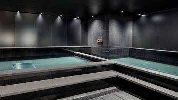 A modern indoor swimming pool with clear water, surrounded by dark walls and steps leading into the pool.