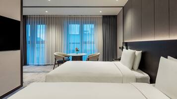 A hotel room with a large bed, a round table with two chairs, a TV, and a view of the city through sheer curtains.