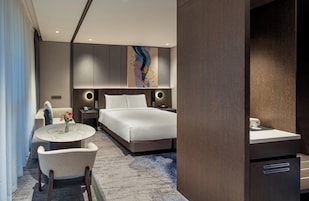 Una habitación de hotel moderna con una cama, mesitas de noche, una pequeña mesa redonda con un jarrón de flores y vistas al pasillo.