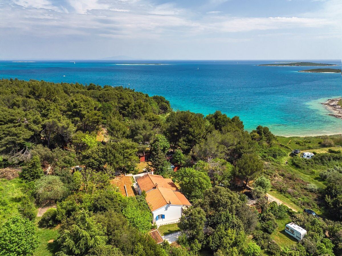 A coastal property with a house surrounded by dense greenery, a clear view of the sea, and a small boat docked near the shore.